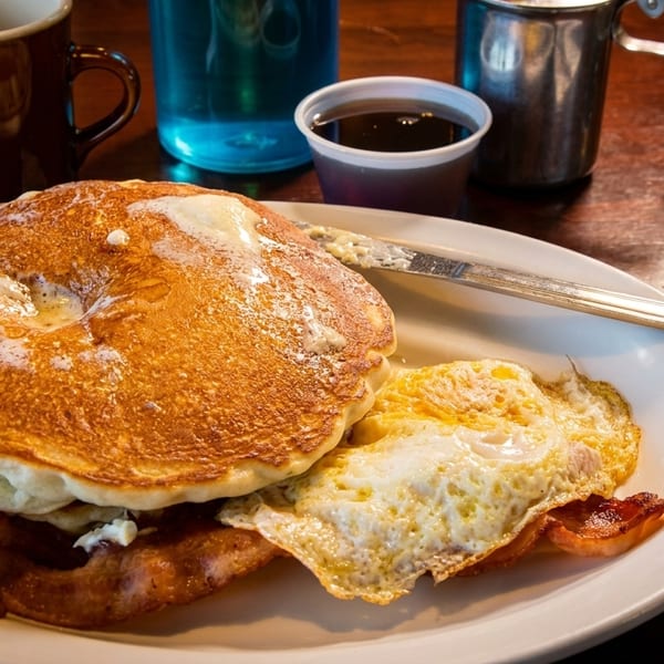 A breakfast plate with pancakes, fried eggs, bacon, and a side of syrup on a wooden table.