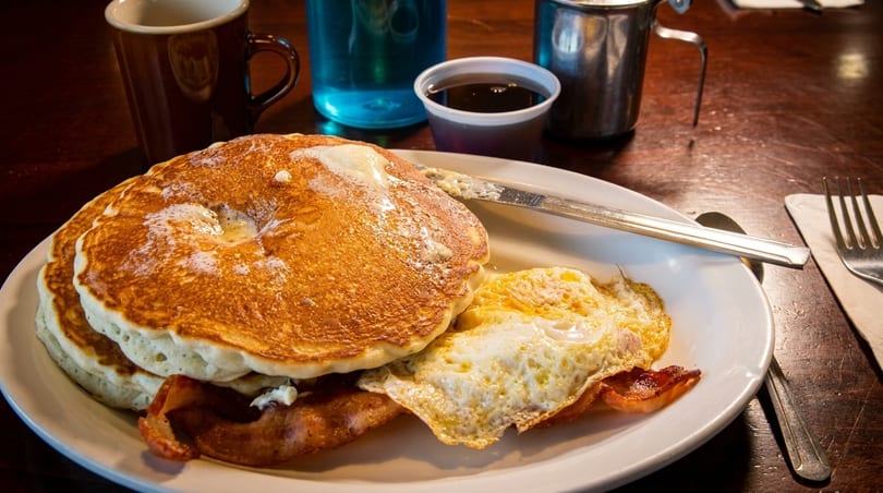 A breakfast plate with pancakes, fried eggs, bacon, and a side of syrup on a wooden table.