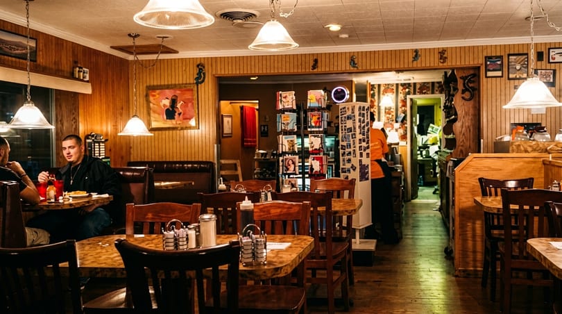Cozy diner interior with wood paneling, hanging lights, and patrons dining at tables and booths.