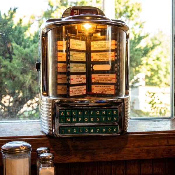 A vintage chrome jukebox with glowing lights sits on a wooden windowsill, with trees visible outside.