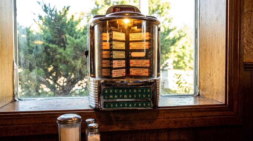 A vintage chrome jukebox with glowing lights sits on a wooden windowsill, with trees visible outside.