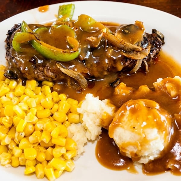 Salisbury steak with gravy, peppers, onions, mushrooms, mashed potatoes, and corn on a white plate.