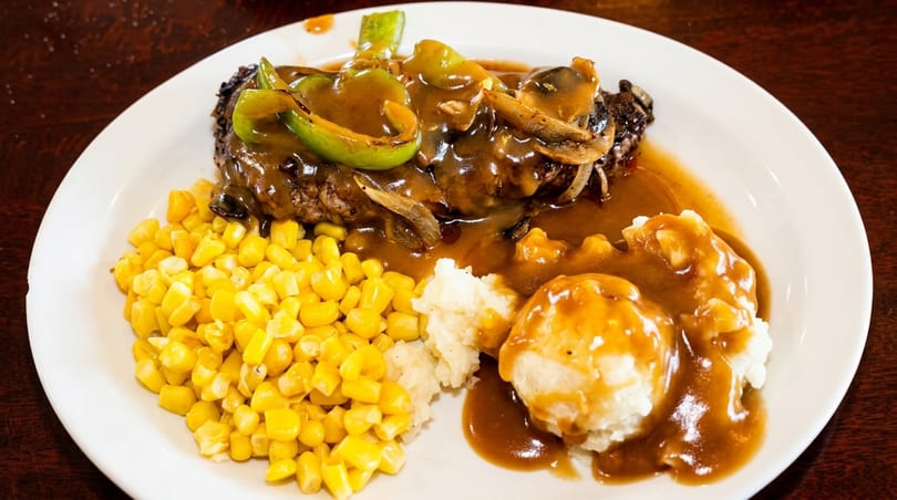 Salisbury steak with gravy, peppers, onions, mushrooms, mashed potatoes, and corn on a white plate.