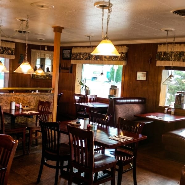 Cozy restaurant interior with wooden tables, chairs, booths, and pendant lights, seen through large windows.