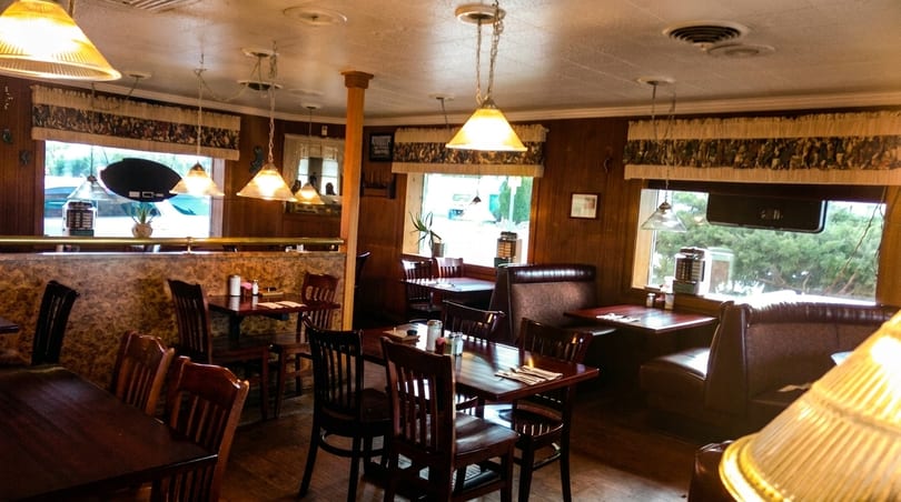 Cozy restaurant interior with wooden tables, chairs, booths, and pendant lights, seen through large windows.