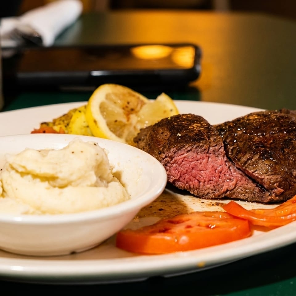 A medium-rare steak, mashed potatoes, sliced tomatoes, and lemon on a white plate.