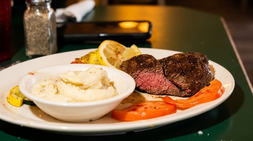 A medium-rare steak, mashed potatoes, sliced tomatoes, and lemon on a white plate.