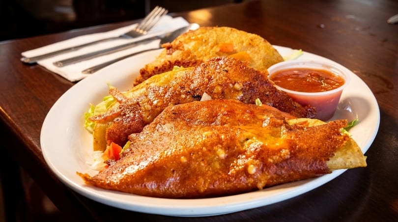 Three crispy tacos with lettuce, tomato, and a side of salsa on a white plate.
